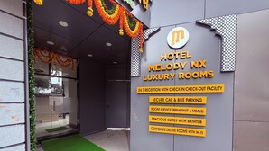 Entrada de hotel con un cartel que anuncia 'Hotel Melody NX Luxury Rooms', destacando servicios como recepción las 24 horas, estacionamiento seguro, servicio a la habitación, amplios suites con bañera y habitaciones corporativas de lujo con Wi-Fi.