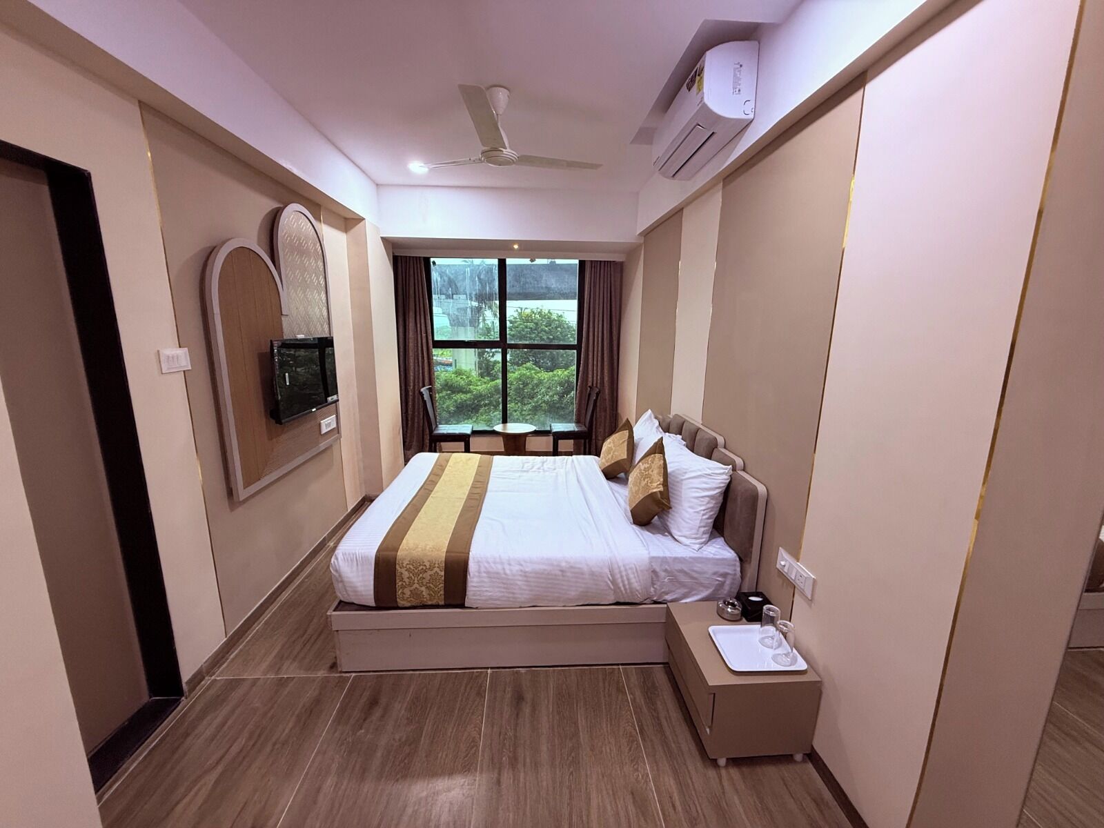 A hotel room with a bed, bedside table, TV, air conditioning unit, and a window with a view.