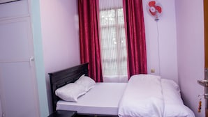 A bedroom with a bed, white bedding, a nightstand, a wall-mounted fan, and red curtains. - SANAWARI LODGE (Arusha)