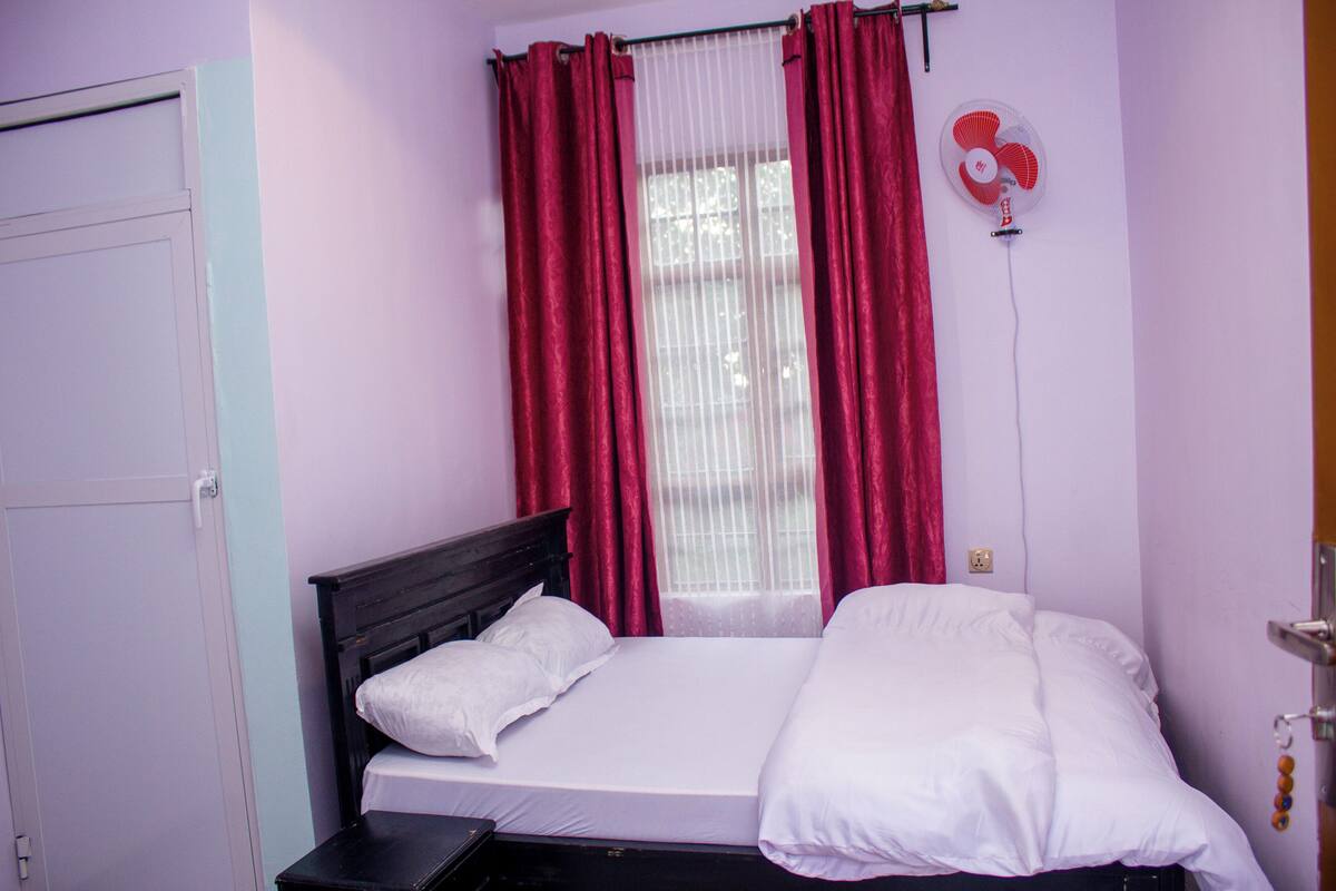 A bedroom with a bed, white bedding, a nightstand, a wall-mounted fan, and red curtains.