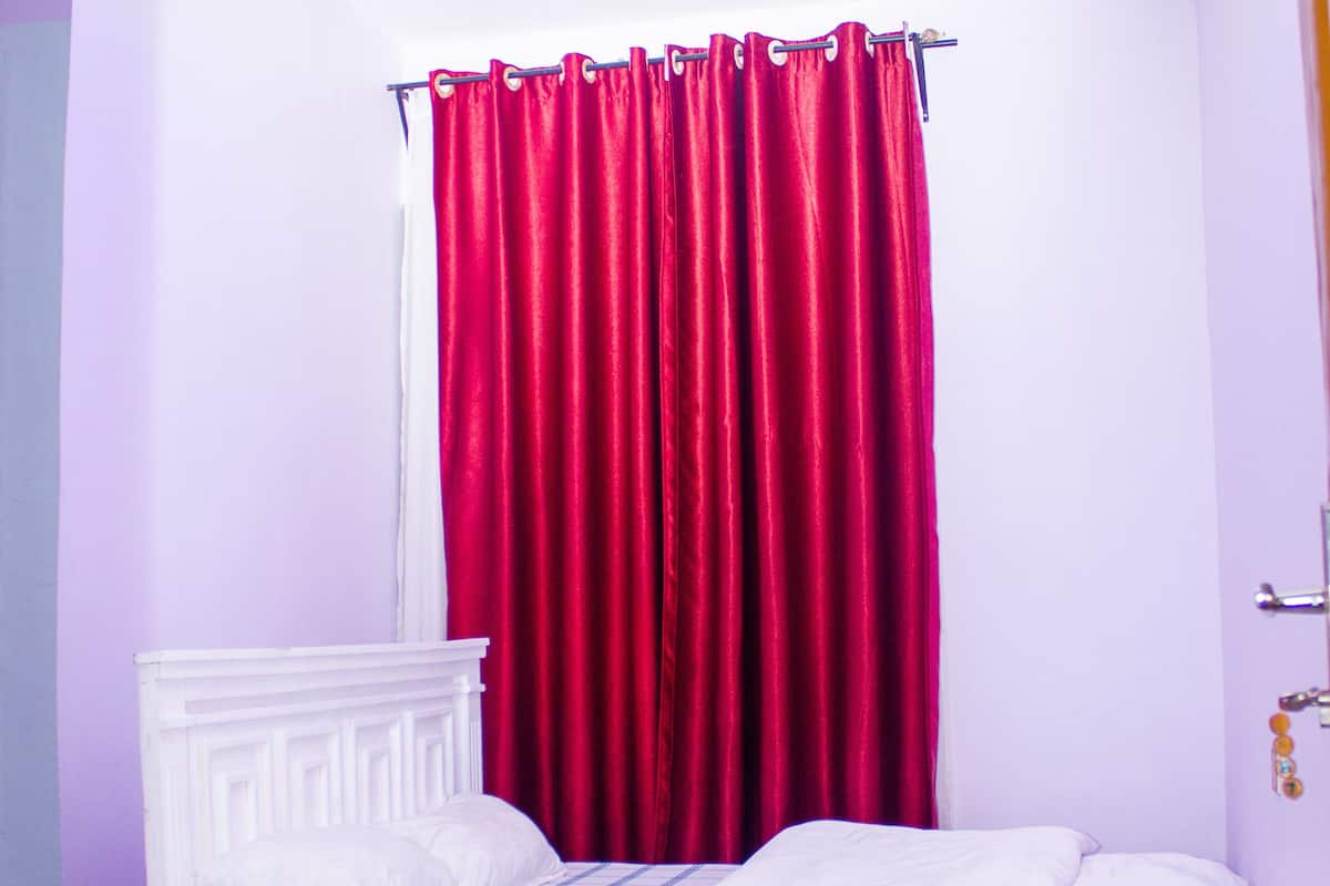 A bedroom with a white bed, a small white bedside table, and a window with red curtains.