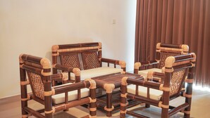 A traditional wooden sofa set with cushioned seats and backrests, positioned in a well-lit room with a plain wall and a window with curtains. - The Raka Villa Amed (Karangasem)