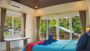 A bedroom with a large bed, a wooden bench, and a view of a lush garden through large windows. - The Raka Villa Amed (Karangasem)