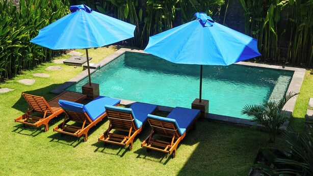 A rectangular swimming pool with a tiled edge, surrounded by a grassy area with stepping stones. There are four wooden lounge chairs with blue cushions and two blue umbrellas providing shade. - The Raka Villa Amed (Karangasem)