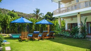 A poolside area with lounge chairs and umbrellas, surrounded by lush greenery and a two-story building with a balcony. - The Raka Villa Amed (Karangasem)