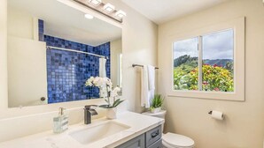Un baño con lavamanos blanco, un espejo, zona de ducha con azulejos azules, inodoro y ventana con vistas a zonas verdes.