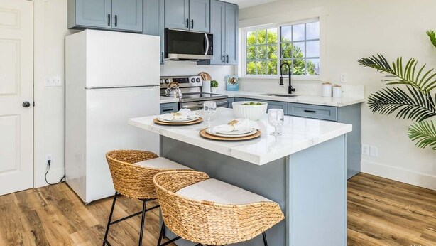 Una cocina moderna con gabinetes grises, un refrigerador blanco, un microondas, una estufa y un lavamanos. Hay dos taburetes de mimbre y una isla grande con encimera blanca.