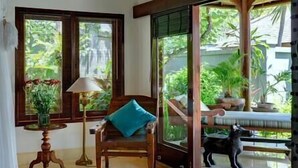 Una habitación con una silla de madera, una mesa con lámpara y vistas a la vegetación exterior.