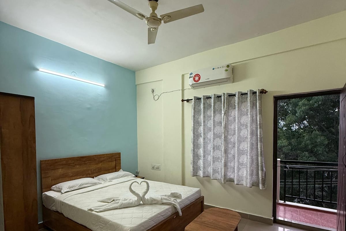 A bedroom with a bed, a ceiling fan, an air conditioning unit, a window with curtains, a small wooden table, and a balcony with a railing.