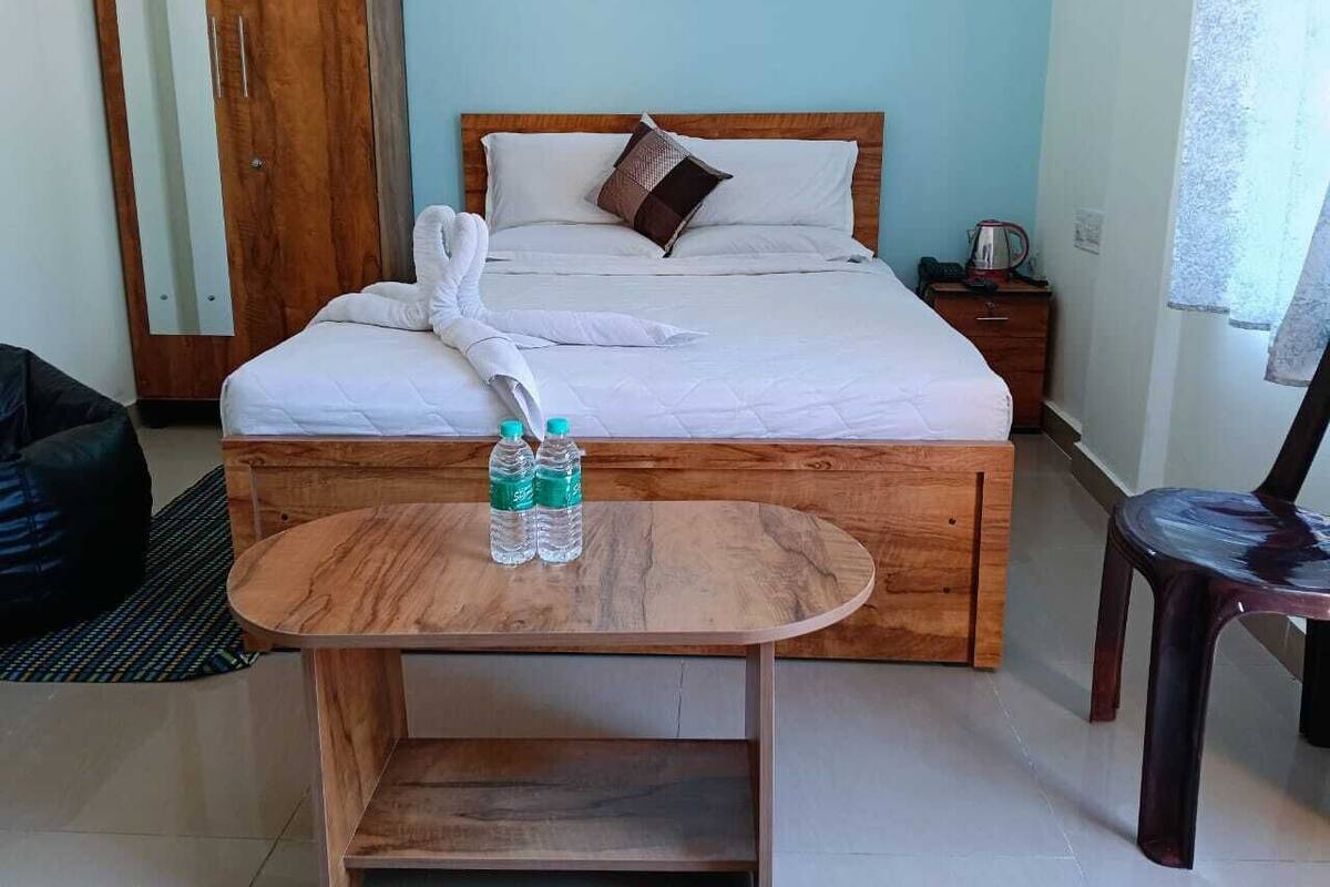 A hotel room with a bed, a wooden nightstand, a small table with water bottles, a black bean bag, and a chair.
