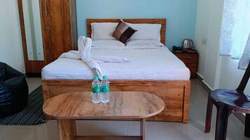 A hotel room with a bed, a wooden nightstand, a small table with water bottles, a black bean bag, and a chair.