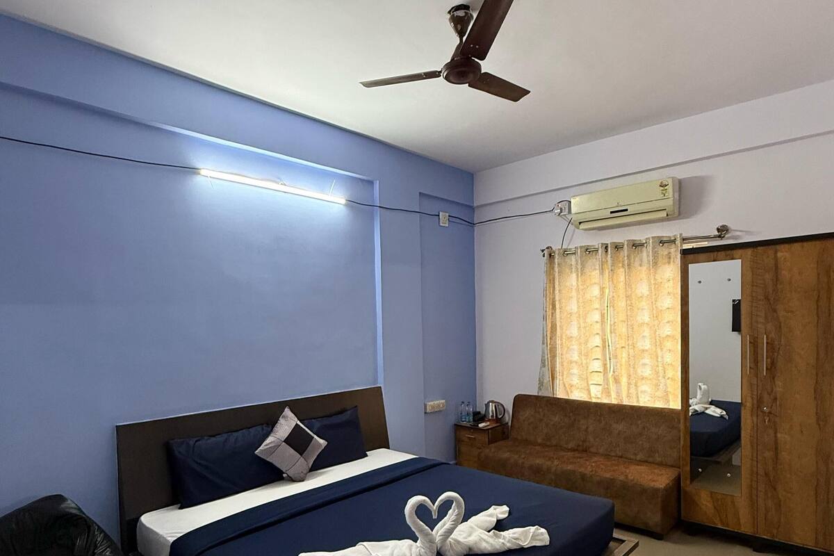 A hotel room with a bed, a ceiling fan, an air conditioning unit, a mirror, and a wooden wardrobe.