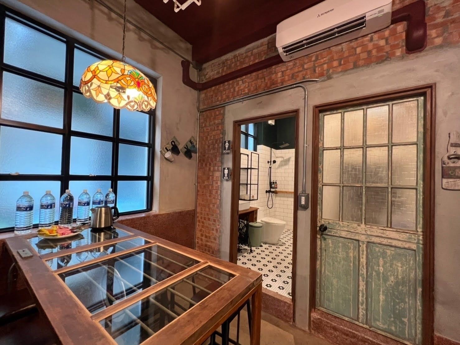 A room with a glass table, water bottles, a decorative light fixture, and a door leading to a shower area.