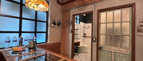 A room with a glass table, water bottles, a decorative light fixture, and a door leading to a shower area.