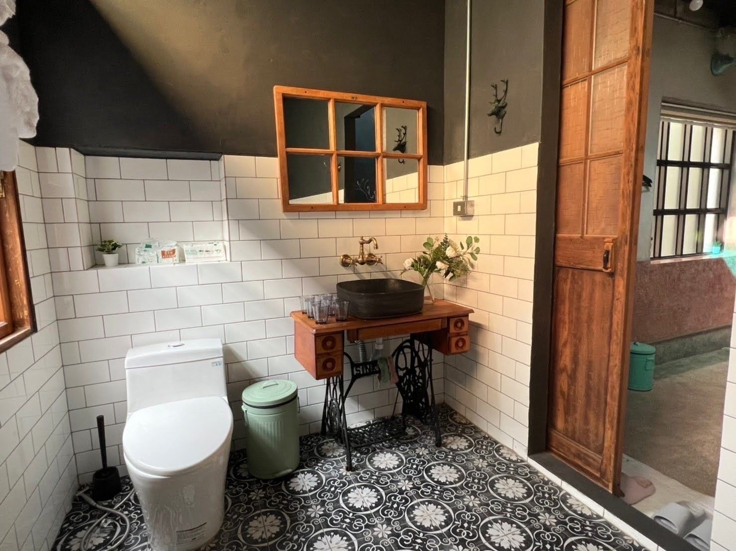 A compact bathroom with white tiled walls, a black and white patterned floor, a wooden vanity with a mirror, a sink, and a toilet.