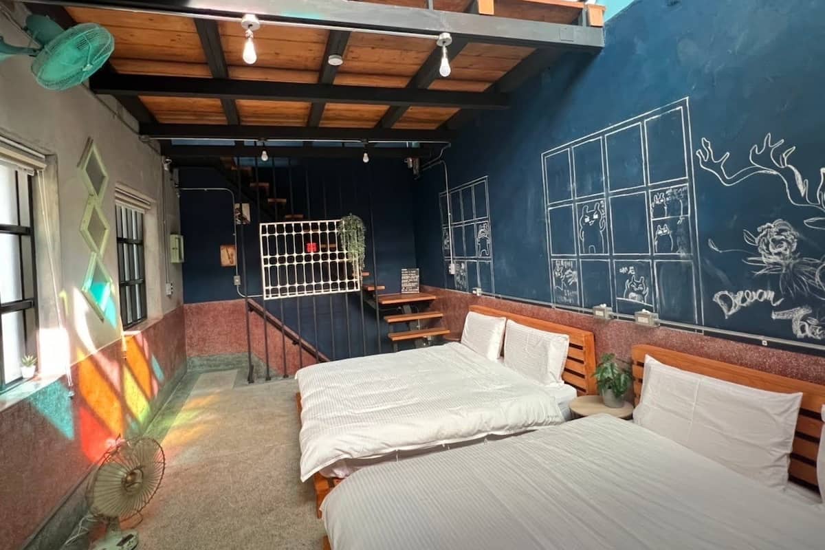Una habitación con dos camas, una mesita, un ventanal y una pared con pizarra y dibujos.