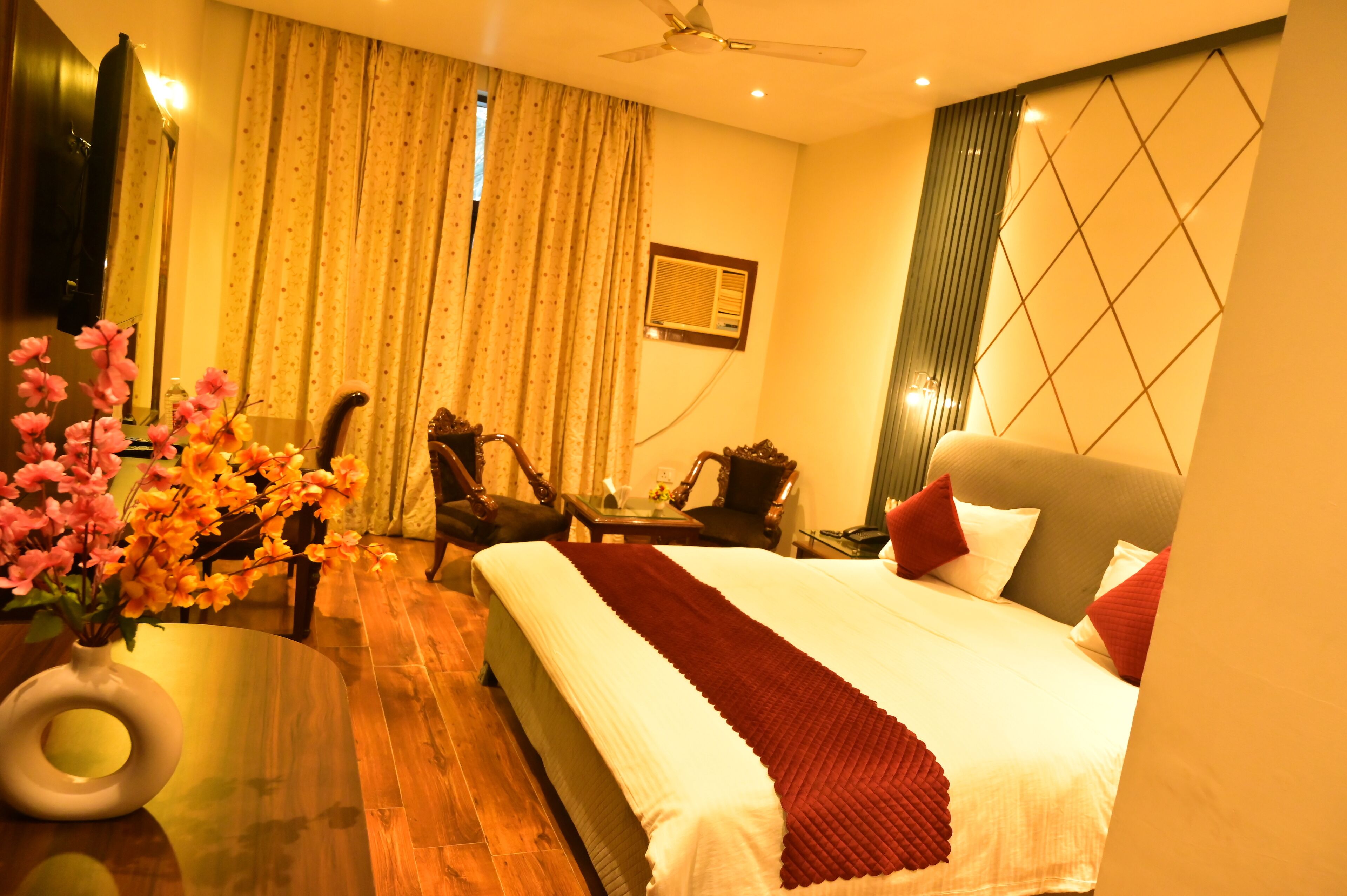 A hotel room with a large bed, a desk with a chair, a small table, a vase with flowers, and a ceiling fan.