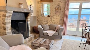 A cozy living room with a stone fireplace, a comfortable sofa, and a view through a large window. - Holiday home: Gîte du Levant (Aiglun)