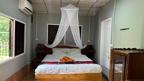 Une chambre avec un lit en bois, du linge de lit blanc, une moustiquaire, une table de chevet en bois, une petite table et une fenêtre donnant sur un paysage verdoyant.