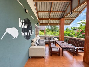 A covered outdoor seating area with a sofa and wooden tables, a wall with a kiwi and animal silhouette, and a view of greenery. - kiwi villas 1 (Vang Vieng)