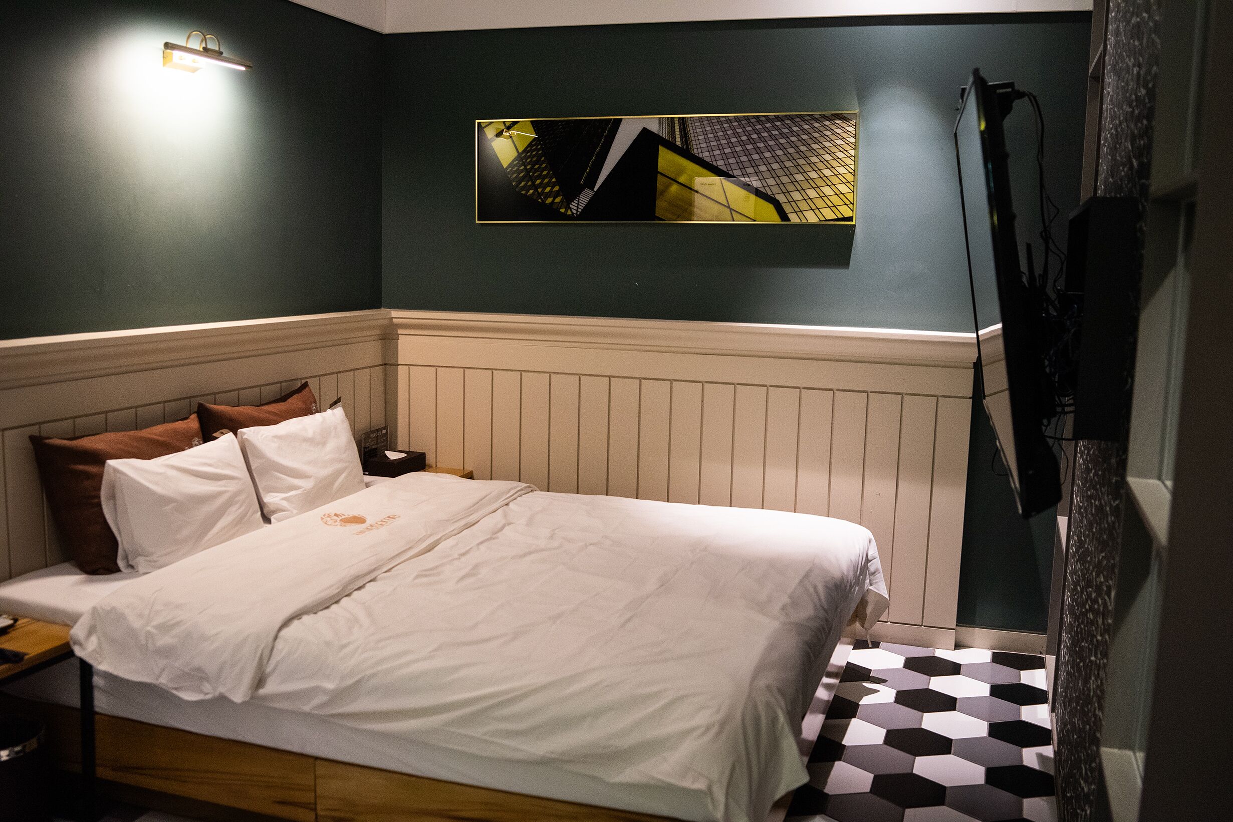 A bedroom with a bed, a nightstand, a wall-mounted TV, and a geometric artwork above the bed.