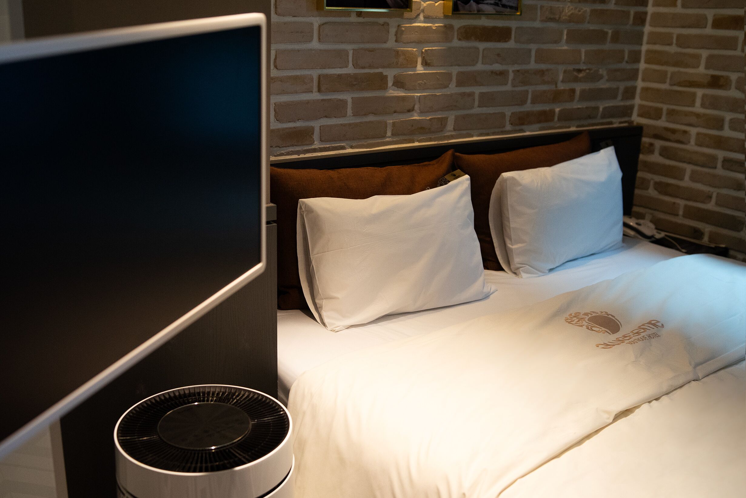 Una habitación de hotel con pared de ladrillo, cama con ropa blanca y un ventilador de pie.