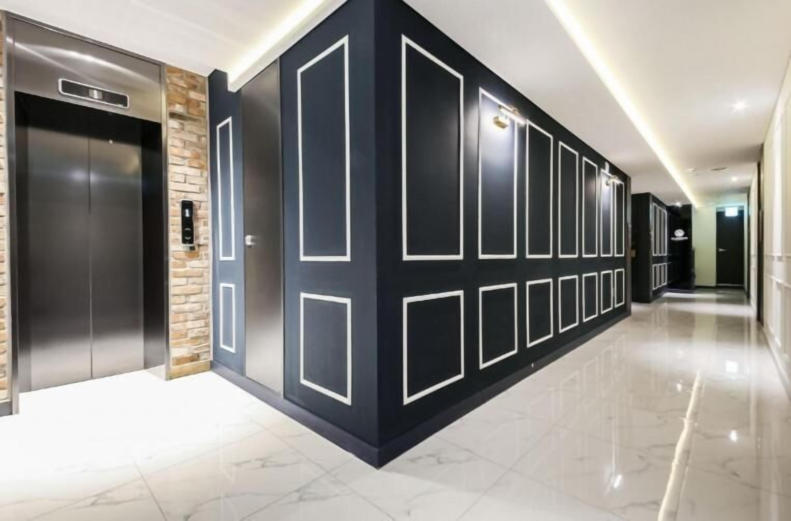 A modern hallway with a polished marble floor, a black accent wall with geometric patterns, and a pair of elevators.