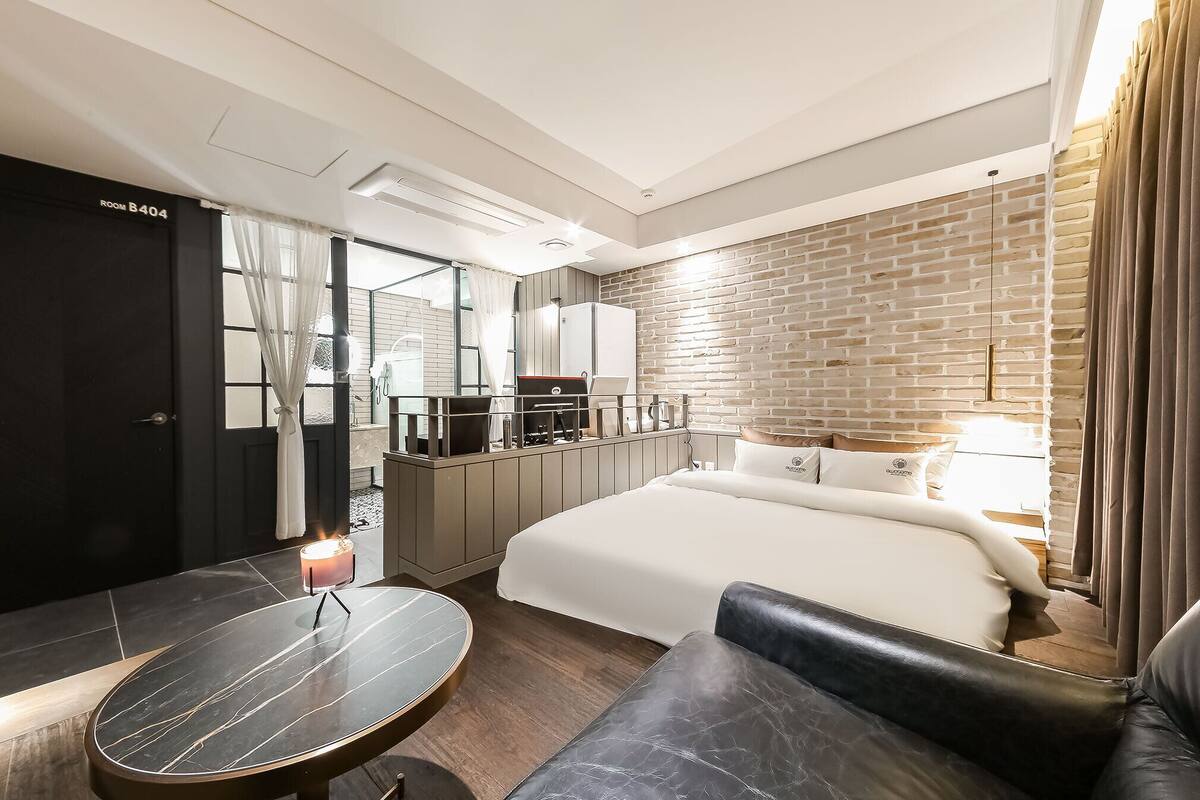 Una habitación de hotel moderna con una cama grande, un escritorio con una computadora portátil, una mesita con una vela y una pared con diseño de ladrillos.