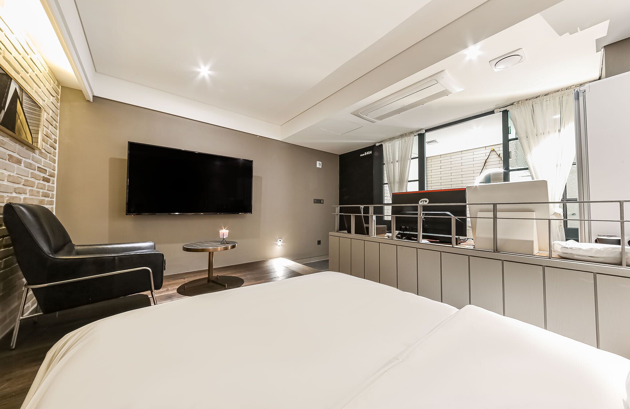 A modern hotel room with a large bed, a flat-screen TV mounted on the wall, a small round table with a candle, and a view of the city through a window.