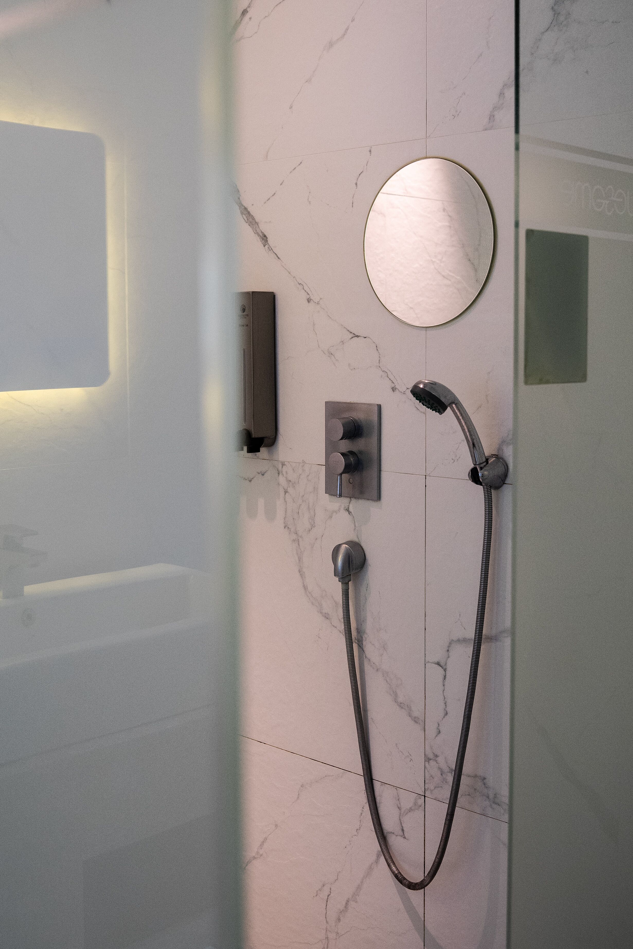 A modern bathroom with a glass shower enclosure, a handheld showerhead, a wall-mounted control panel, and a round mirror.