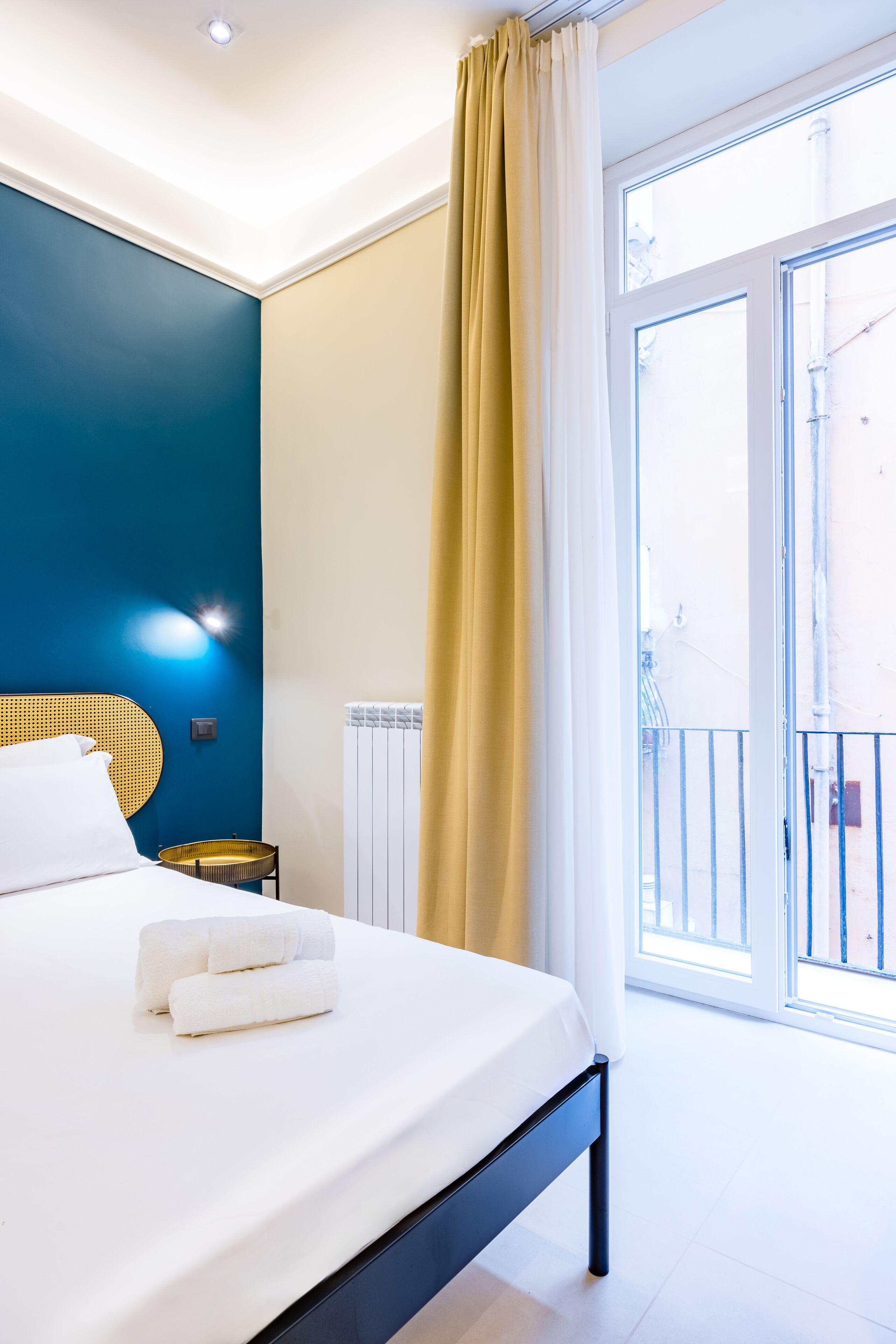 A bedroom with a bed, a bedside table, a wall-mounted light, and a view of the outside.