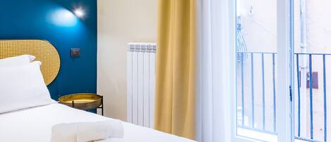 A bedroom with a bed, a bedside table, a wall-mounted light, and a view of the outside.