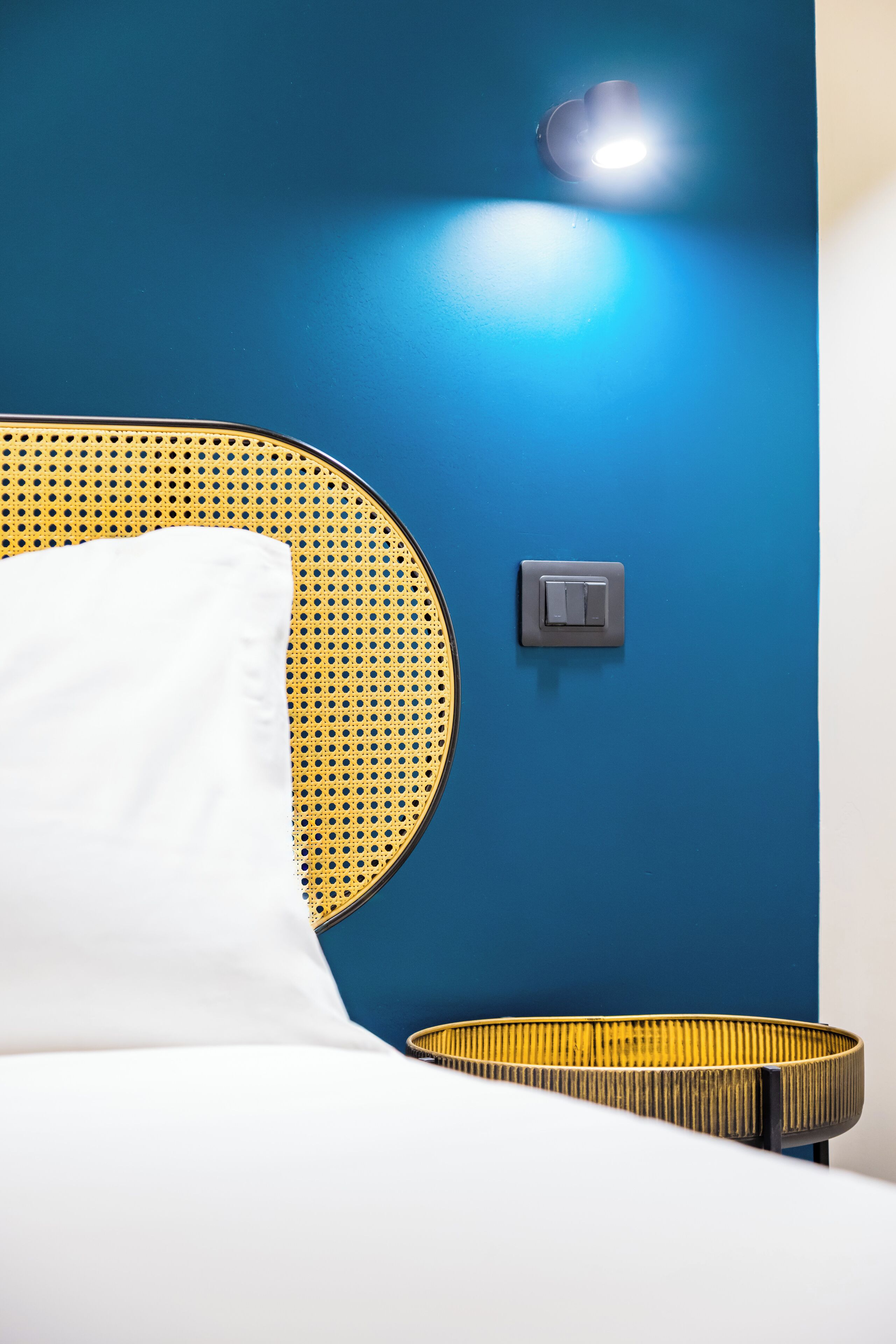A bed with a white pillow and a circular, yellow and black headboard. A light switch and a wall-mounted light are visible on a blue wall.