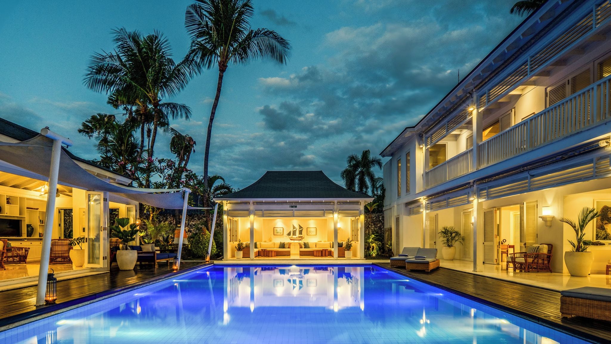 A pool area with lounge chairs and a covered seating area, surrounded by palm trees and buildings.