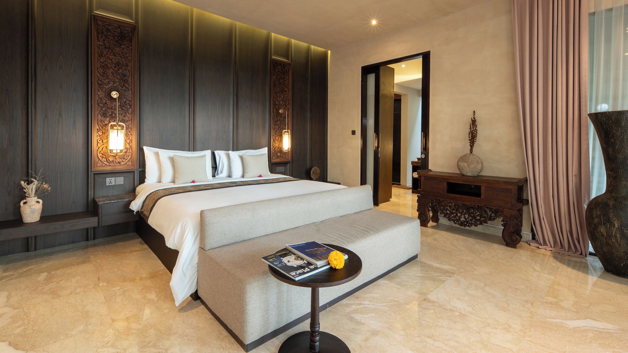 A hotel room with a large bed, bedside lamps, a small round table with a book and a flower, and a decorative vase.