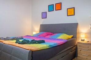 A bed with a rainbow-colored blanket, a gray headboard, and a wooden nightstand with a lamp.