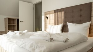 Een net opgemaakt bed met witte lakens en kussens, een houten hoofdbord en een houten plankunit links.