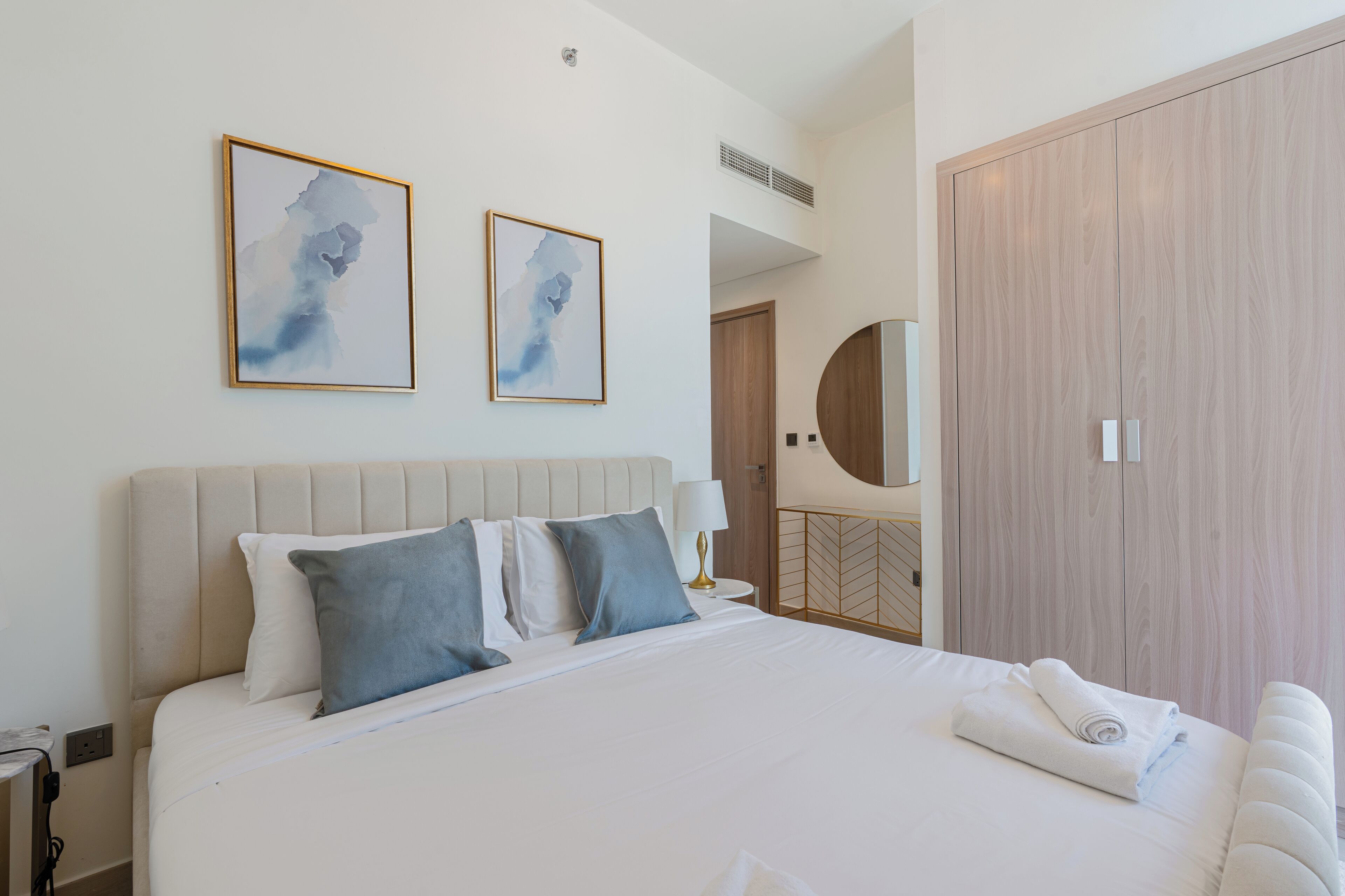 A modern hotel room with a large bed, two framed abstract paintings, a wooden wardrobe, and a round mirror.