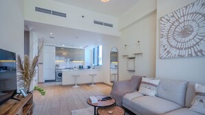 A modern living room with a sofa, a coffee table, and a TV. There is a kitchen in the background with a dining area. - Azizi Riviera Residences - Meydan (Dubai)