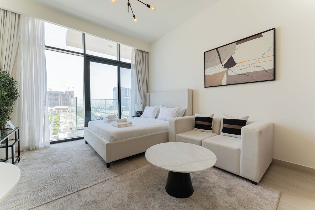 A modern hotel room with a large bed, two sofas, a round coffee table, and a large window offering a city view.