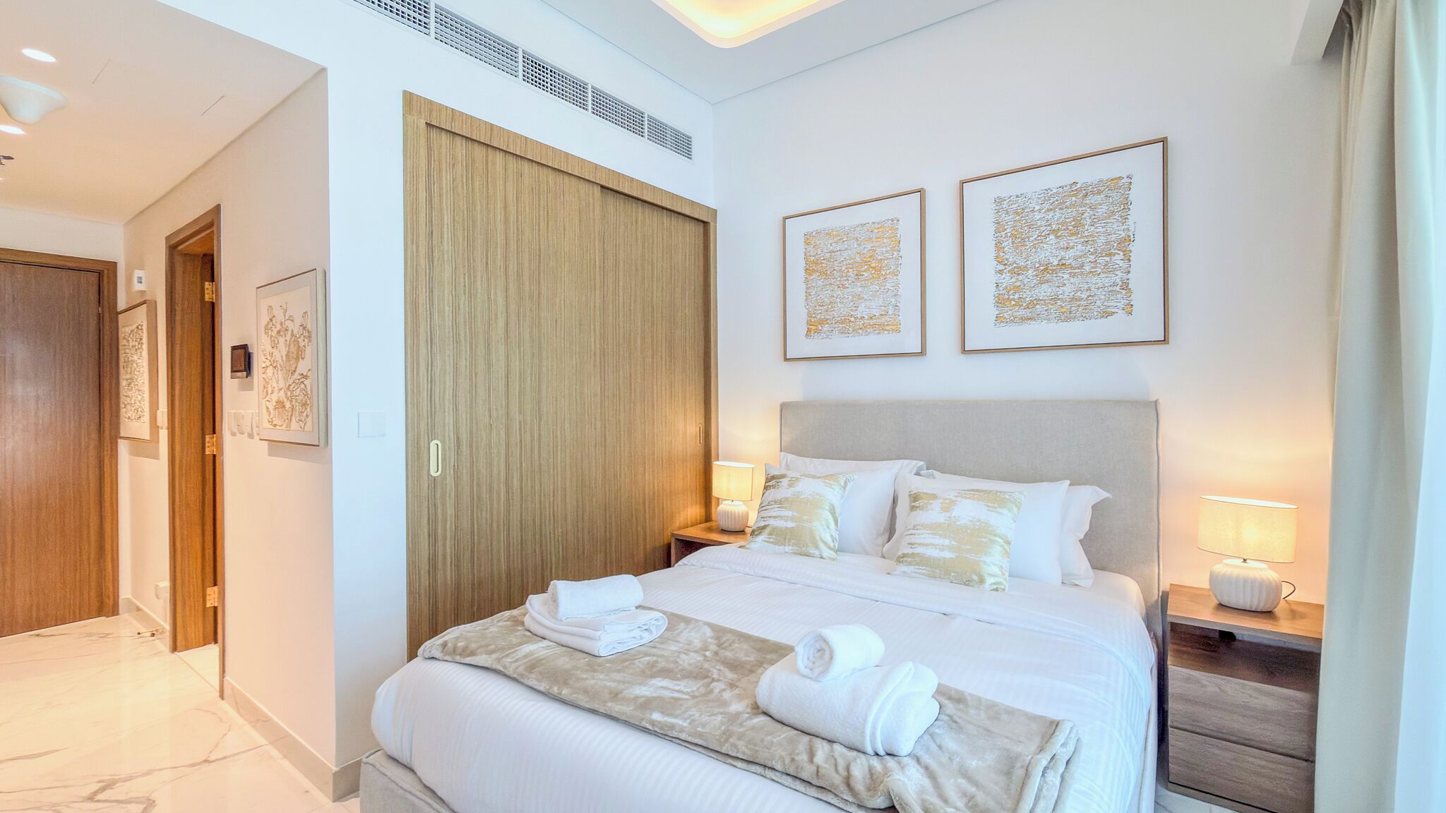 A hotel room with a bed, a wooden wardrobe, two bedside tables with lamps, and two framed artworks on the wall.
