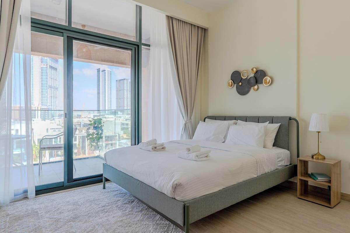 A modern hotel room with a large bed, a nightstand, a lamp, and a view of the city through floor-to-ceiling windows.