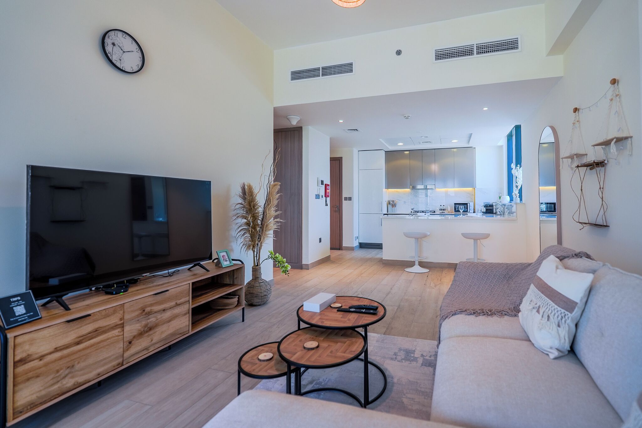 A modern living room with a flat-screen TV, wooden entertainment unit, and a cozy seating area. A kitchen with a central island and pendant lighting is visible in the background.