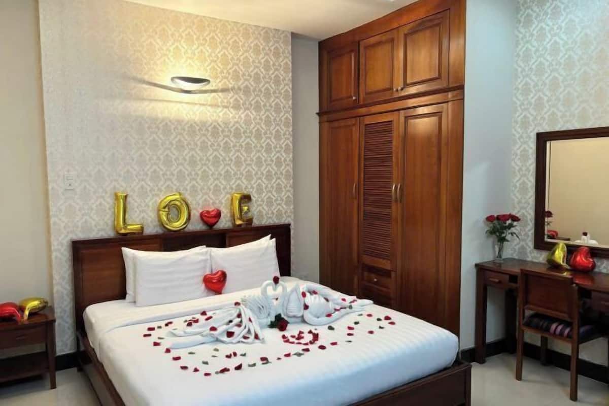 A hotel room with a bed, a wooden wardrobe, a desk with a mirror, and a wall with patterned wallpaper.