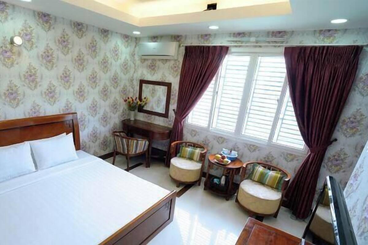A hotel room with a bed, a desk, two chairs, a small table with fruit, and a window with curtains.