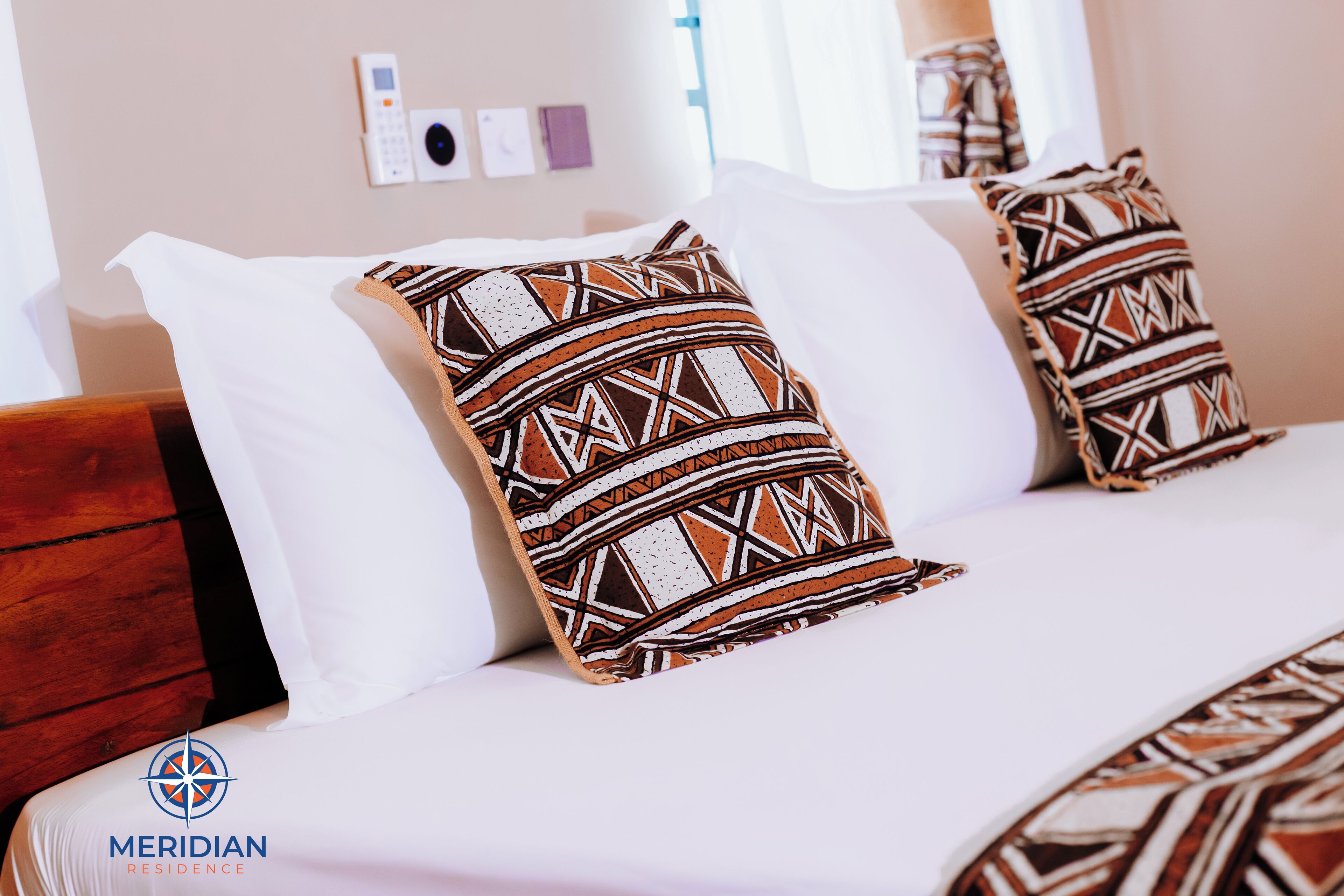 Una habitación de hotel con cama, almohadas estampadas, cabecero de madera, panel de control en la pared y una ventana con cortinas.