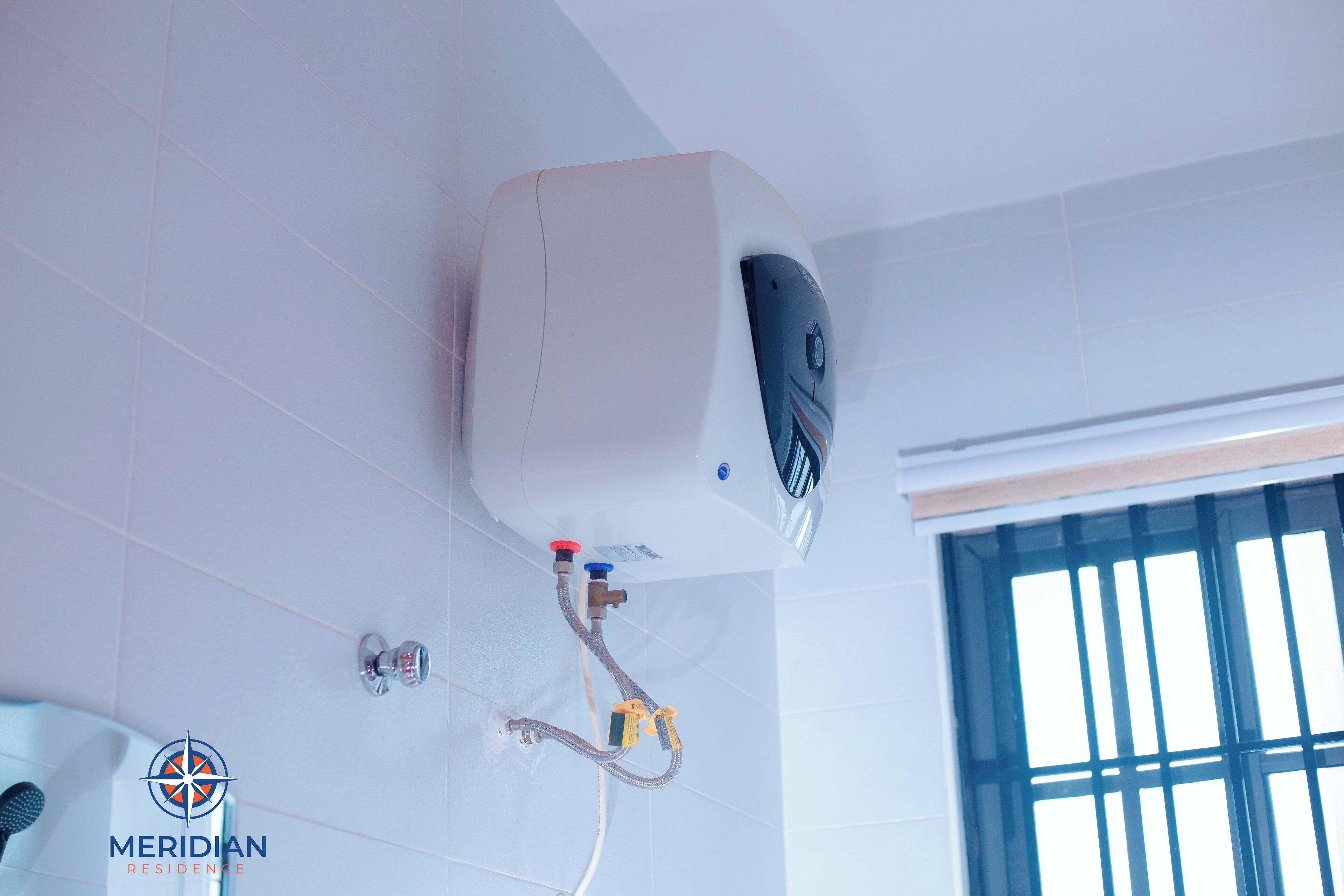 A water heater mounted on a white tiled wall with a window to the right.