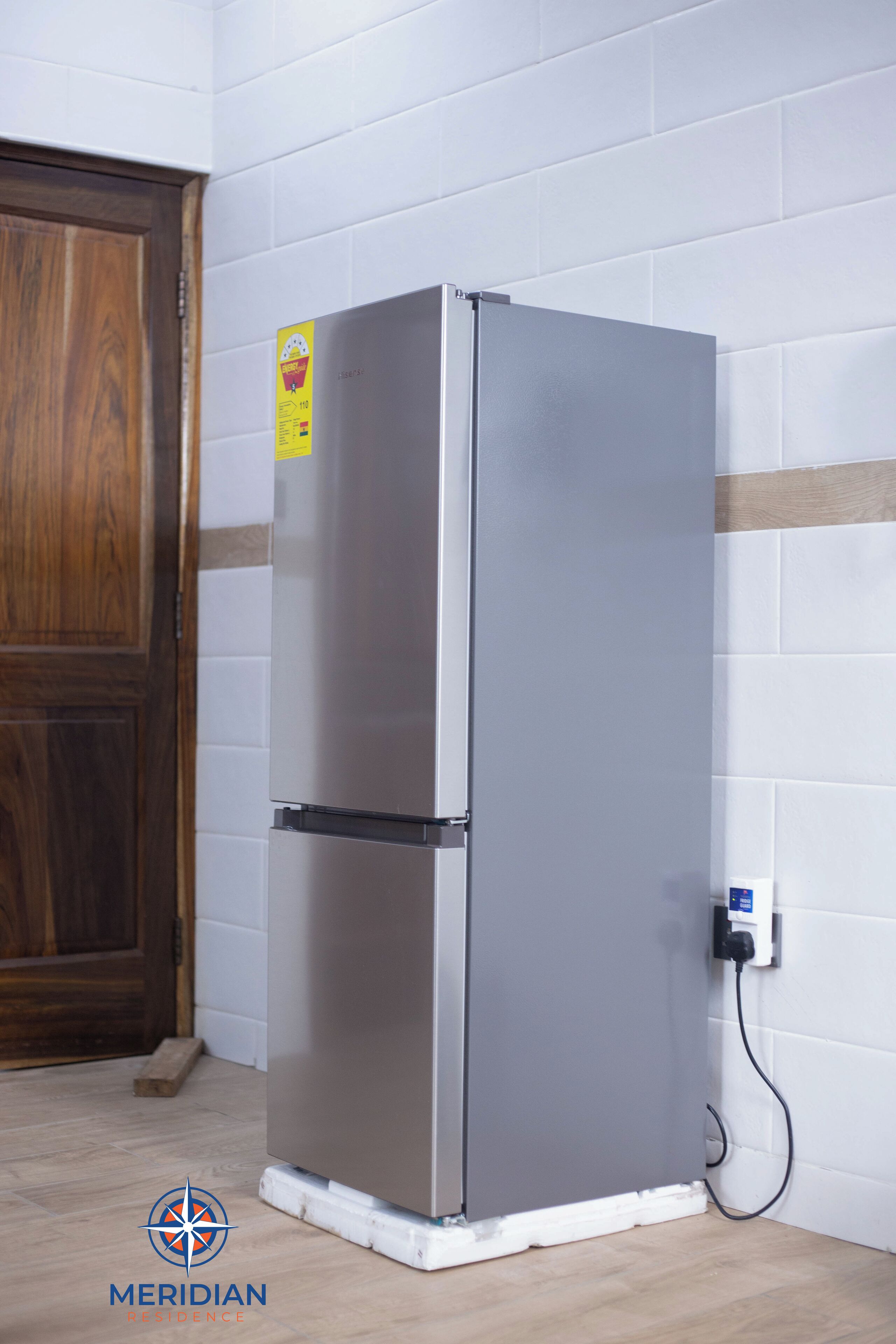A stainless steel refrigerator in a kitchen with white tiled walls and a wooden door.