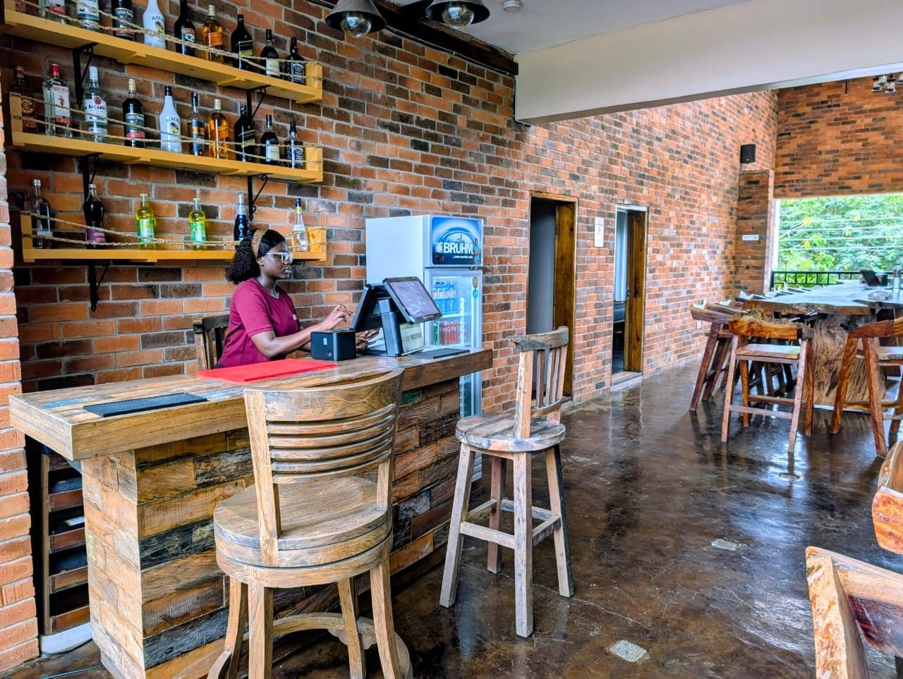 Un espace bar avec un mur en briques, un comptoir en bois, des tabourets et des étagères remplies de bouteilles.