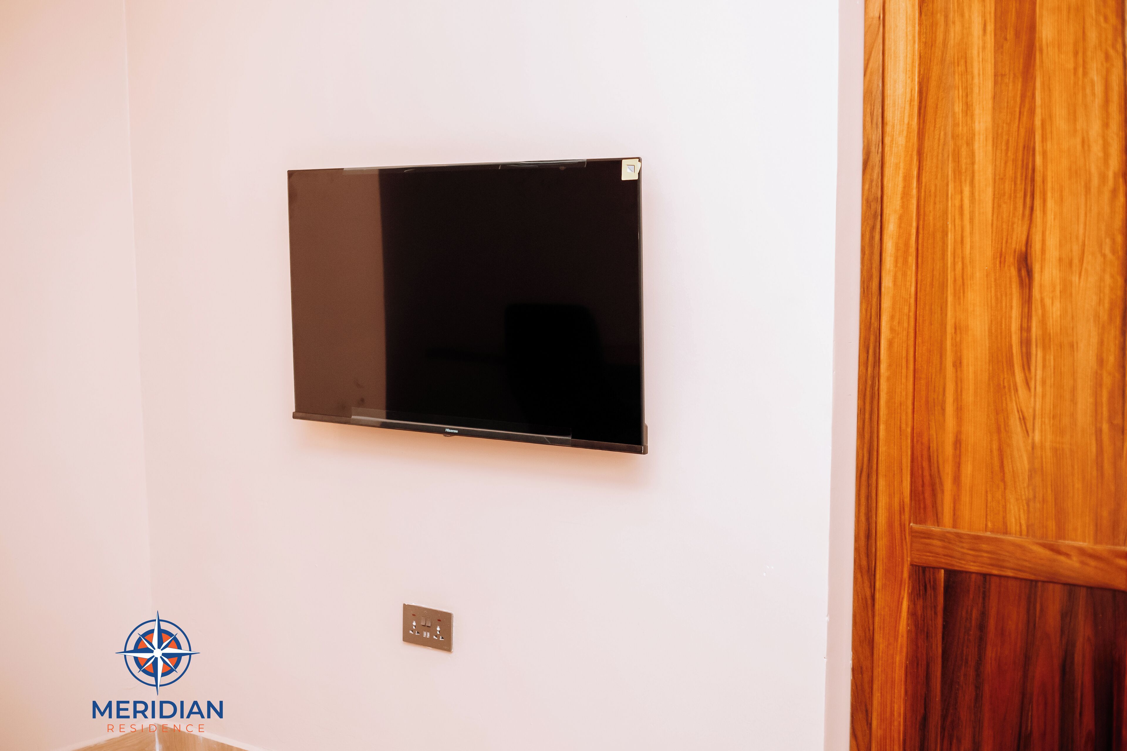 Un téléviseur à écran plat fixé à un mur blanc, une porte en bois sur la droite et le logo de « Meridian Residence » dans le coin inférieur gauche.